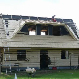 Budowa domu z drewnianych bali z charakterystycznym dachem pokrytym drewnianymi gontami; widoczny pracownik na dachu oraz drabiny opierające się o ściany.